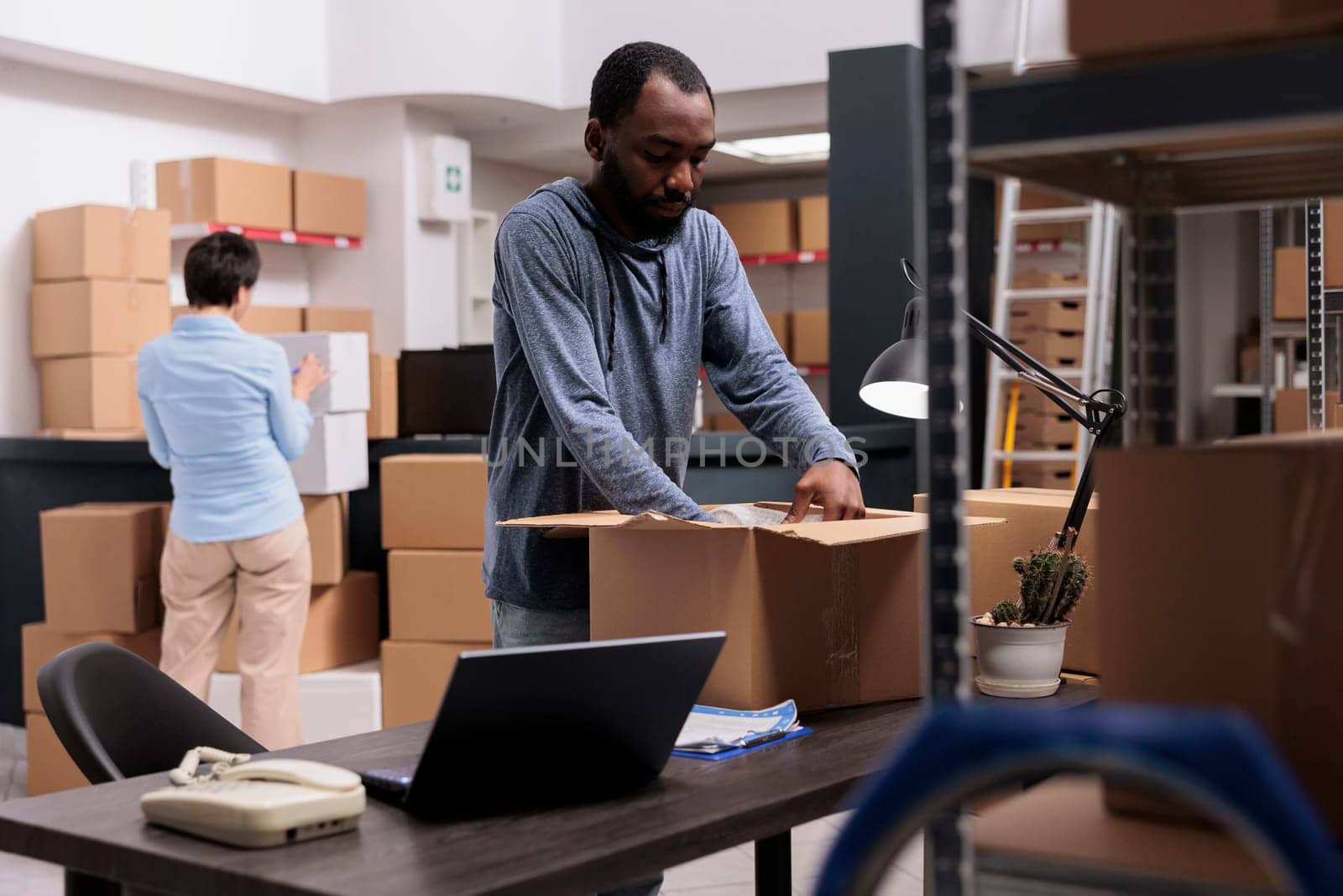 African american employee putting bubble wrap on client order to protect package during transportation, checking shipping detalies on laptop. Diverse team working in warehouse delivery department