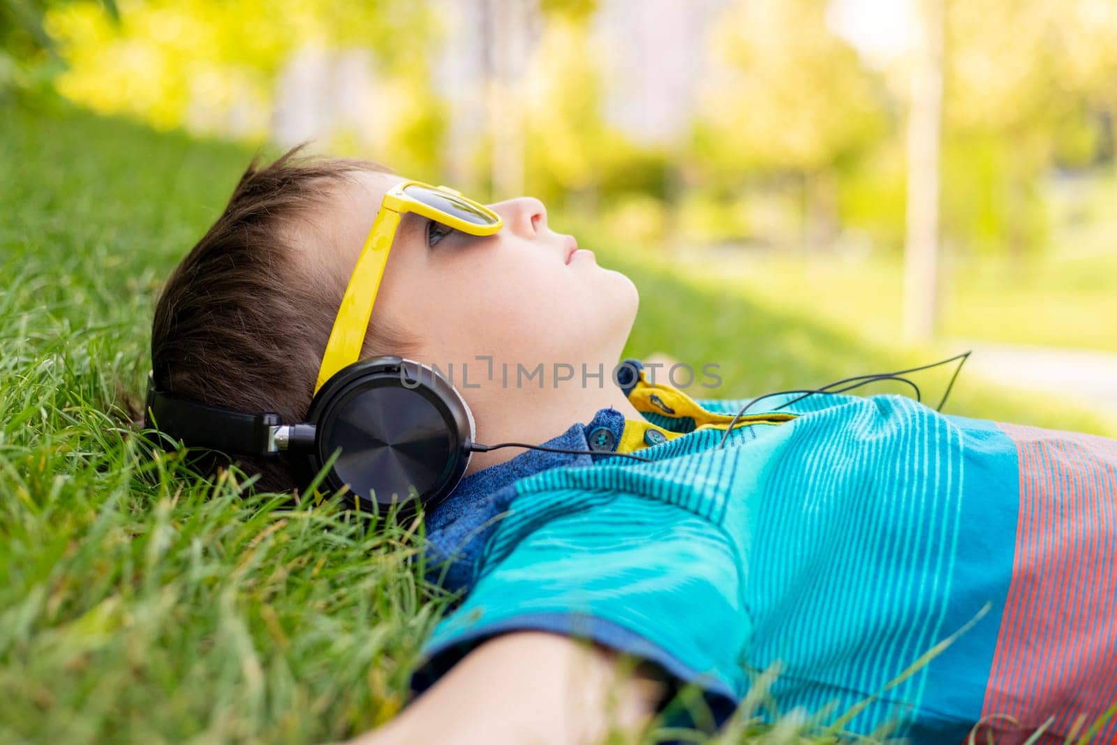 Happy boy in sunglasses lying on the grass and listening to music in headphones on a sunny day.