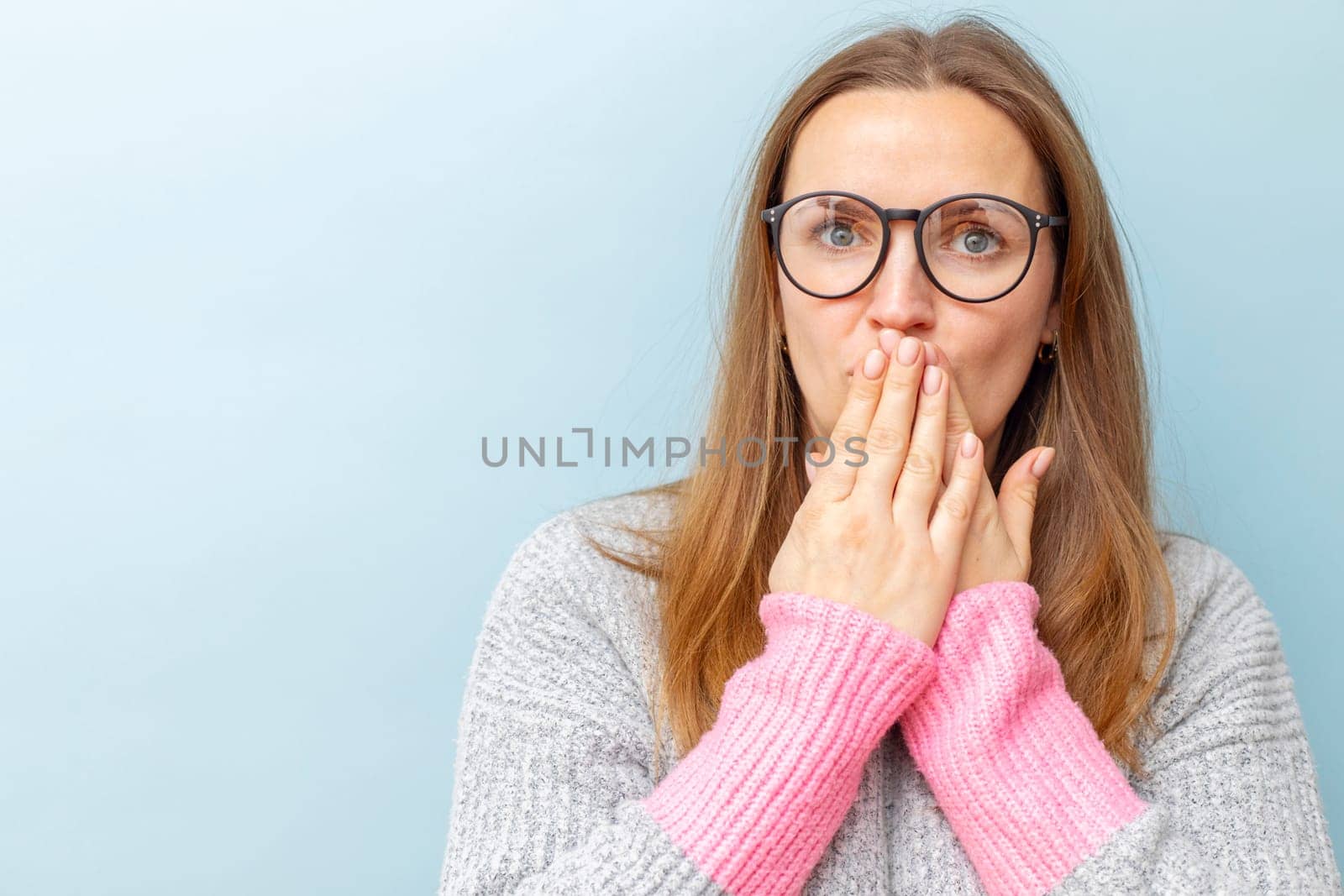 Shocked young woman in eyeglasses covering her mouth with her hands and looking at camera on blue background.