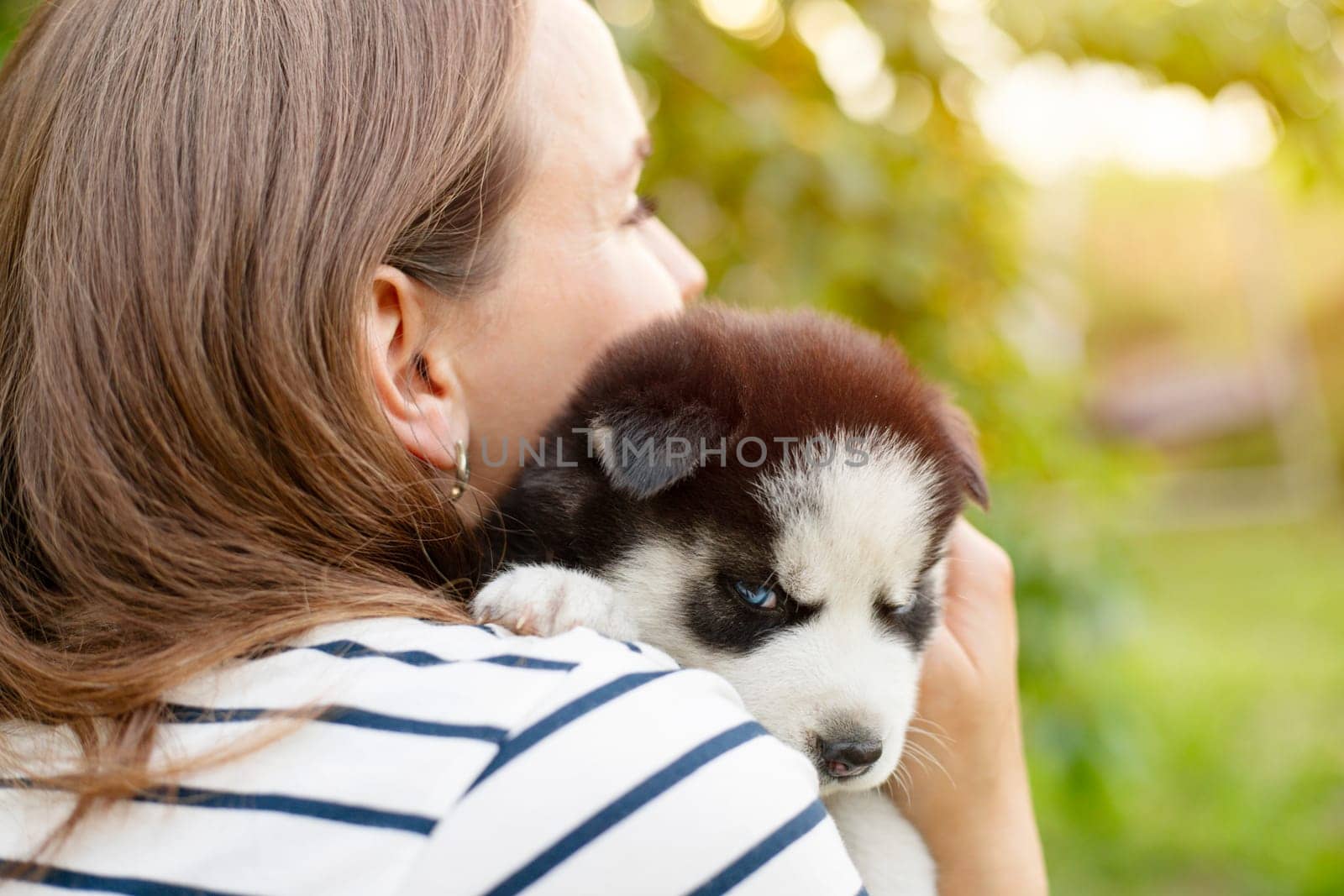 Beautiful woman in a striped t-shirt tenderly hugs a small husky puppy in the park outdoors.