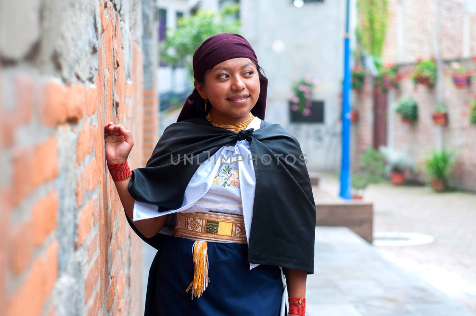 Front view of a confident young Latin woman dressed in traditional clothing from her country standing and leaning her hand against a brick wall looking away outdoors.