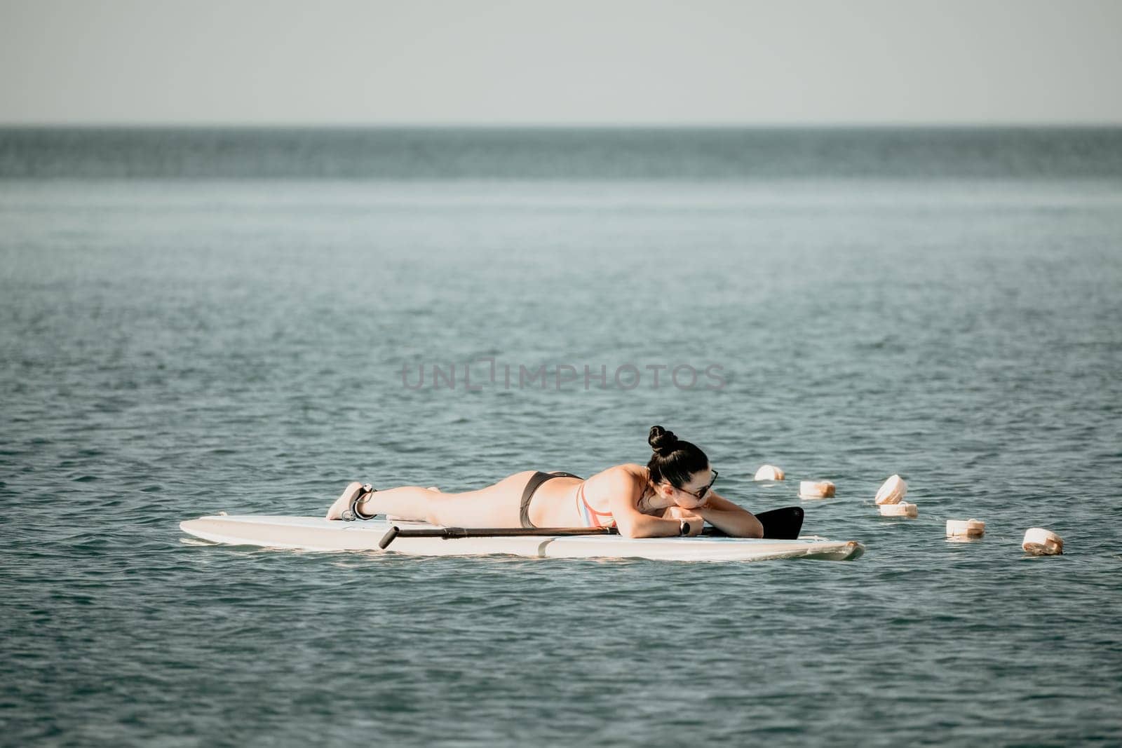 Sea woman sup. Silhouette of happy middle aged tanned woman in rainbow bikini, surfing on SUP board, confident paddling through water surface. Idyllic sunset. Active lifestyle at sea or river. by panophotograph