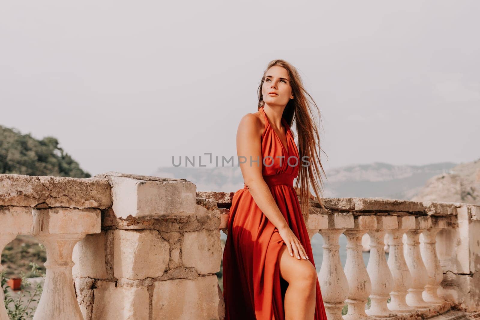 Side view a Young beautiful sensual woman in a red long dress posing on a volcanic rock high above the sea during sunset. Girl on the nature on overcast sky background. Fashion photo