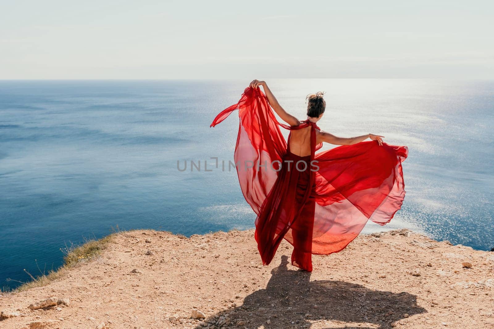 Woman in red dress on sea. Side view a Young beautiful sensual woman in a red long dress posing on a rock high above the sea on sunset. Girl on the nature on blue sky background. Fashion photo. by panophotograph