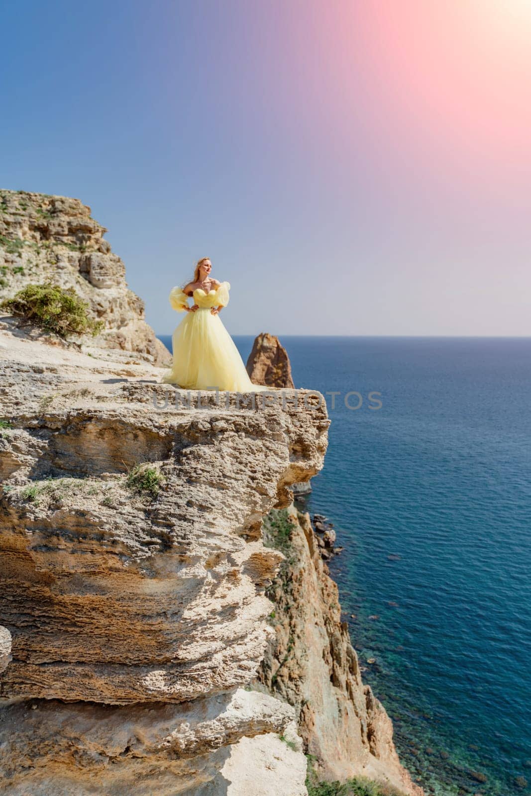 Woman yellow dress sea. Side view Young beautiful sensual woman in yellow long dress posing on a rock high above the sea at sunset. Girl in nature against the blue sky by Matiunina