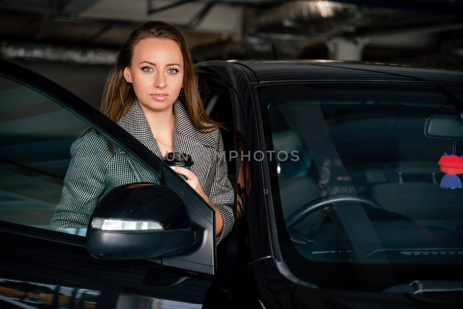 Happy woman car. she stands next to the car in the underground parking. Dressed in a gray coat, holding a glass of coffee in her hands, a black car. by Matiunina