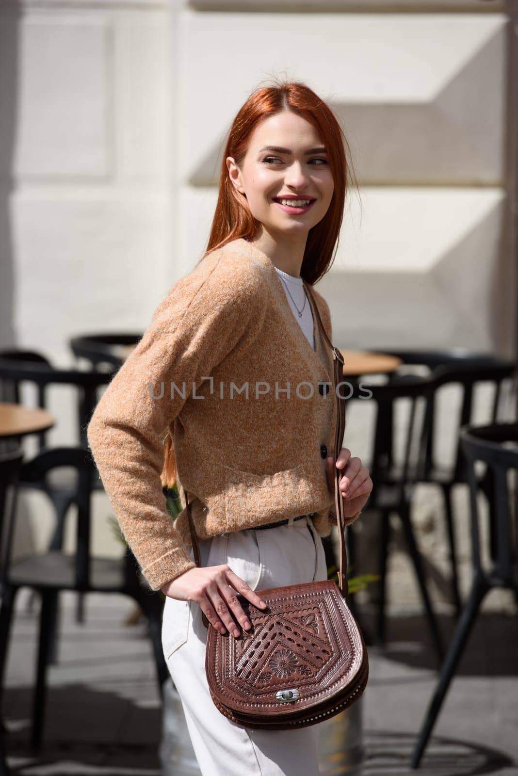 a red-haired girl in white pants and a beige sweater poses outside with a small leather handbag.