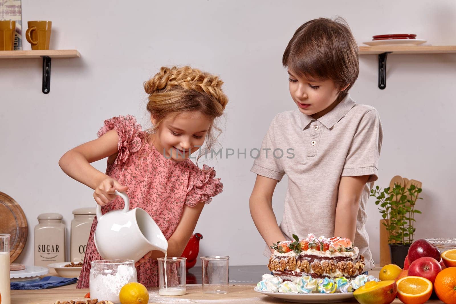 Little brunette boy dressed in a light t-shirt and jeans and a beautiful girl wearing in a pink dress are making a cake at a kitchen, against a white wall with shelves on it. Girl is pouring some milk in a glass.
