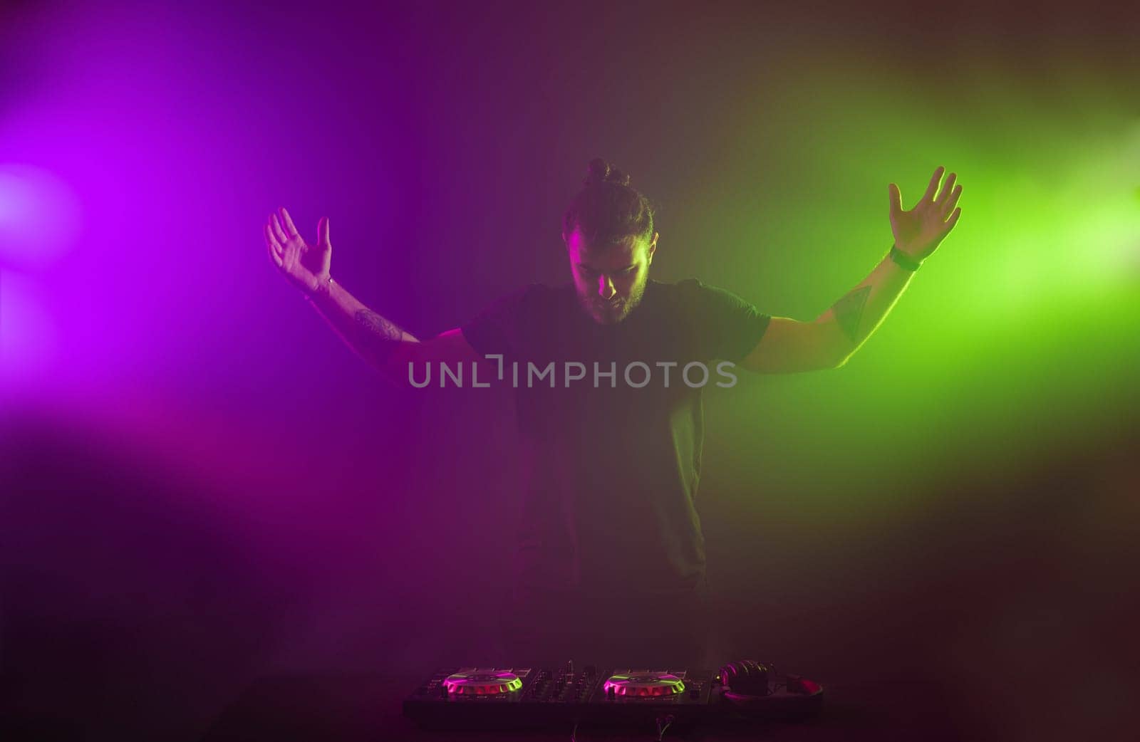 Handsome DJ in a black T-shirt at work mixing sound on her decks at a party or night club with colourful smoke light background