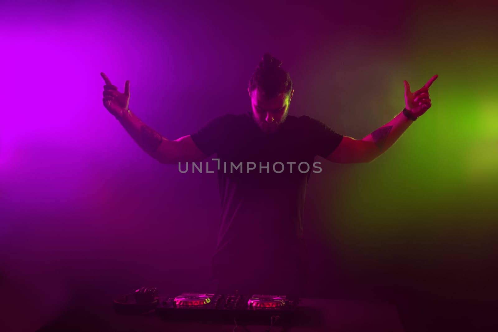 Handsome DJ in a black T-shirt at work mixing sound on her decks at a party or night club with colourful smoke light background