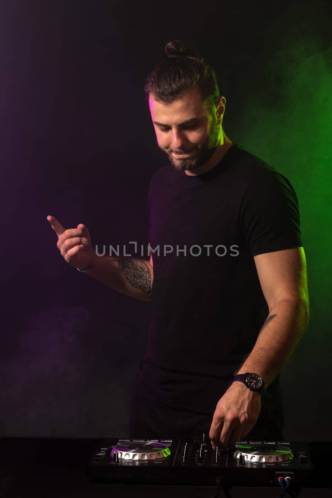 DJ playing music at mixer on colorful foggy background. A fashionable, handsome young man in a black T-shirt is behind the DJ's console. Studio shot