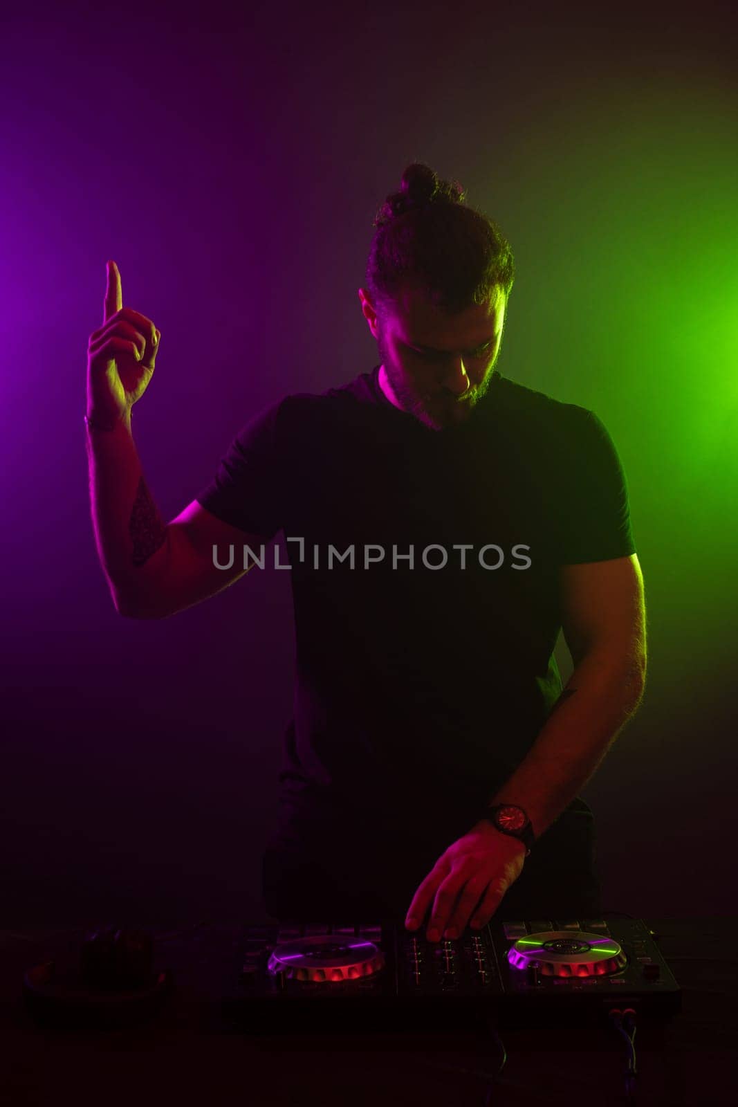 DJ playing music at mixer on colorful foggy background. A fashionable, handsome young man in a black T-shirt is behind the DJ's console. Studio shot