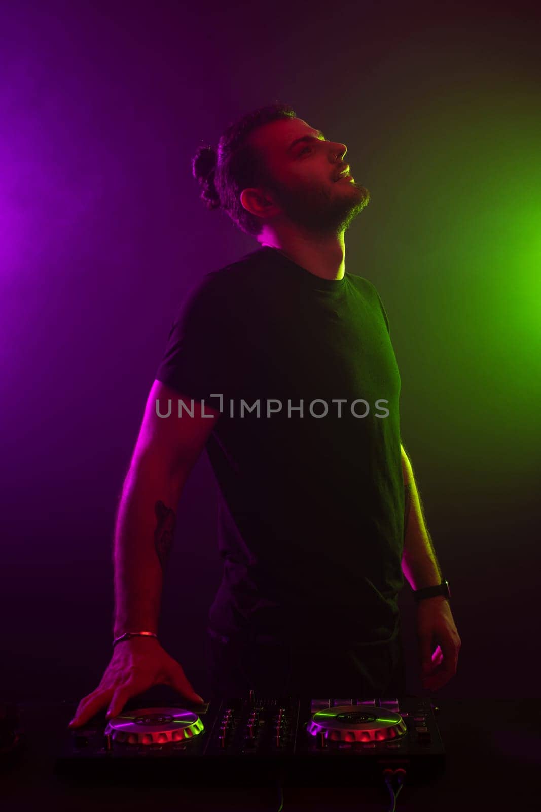 DJ playing music at mixer on colorful foggy background. A fashionable, handsome young man in a black T-shirt is behind the DJ's console. Studio shot