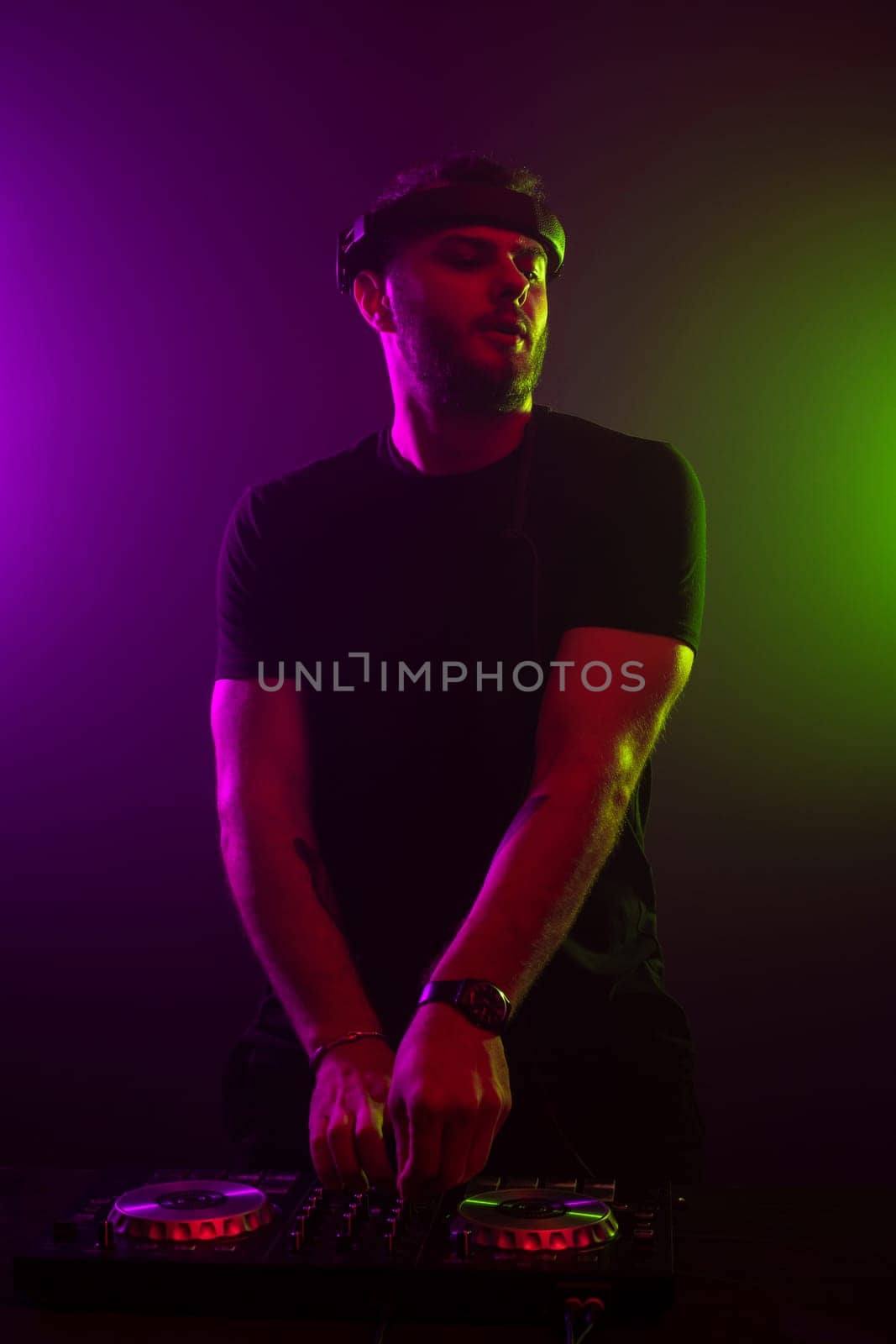 DJ playing music at mixer on colorful foggy background. A fashionable, handsome young man in a black T-shirt is behind the DJ's console. Studio shot