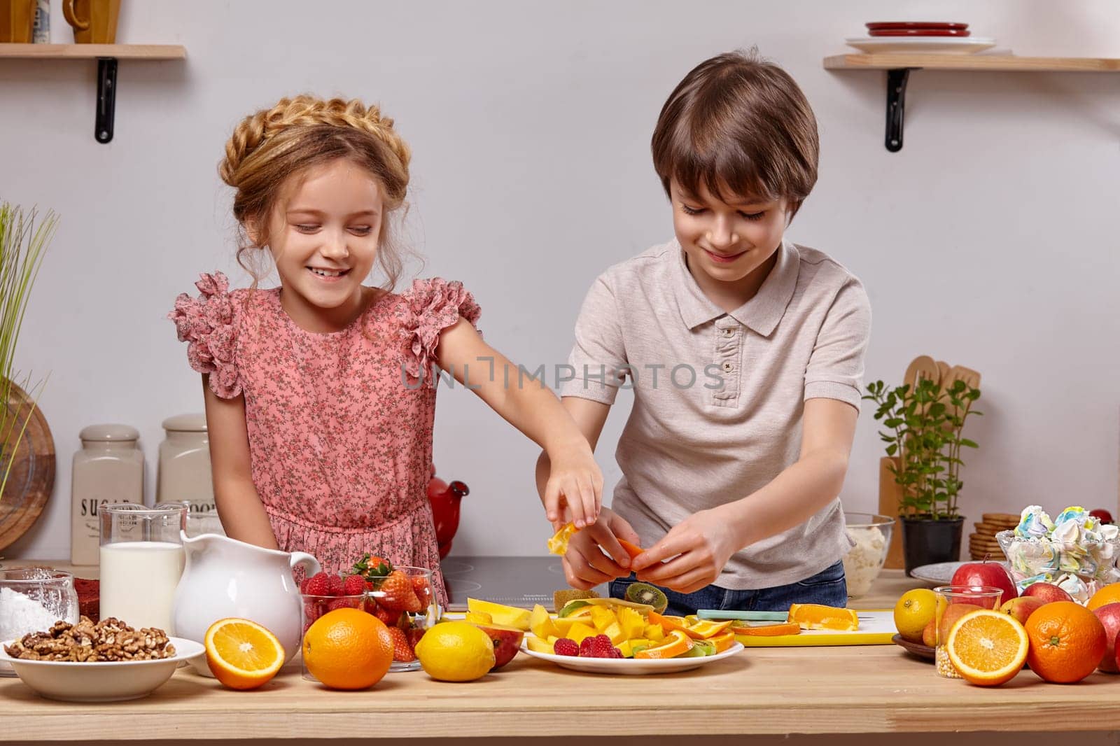 Cute cook couple. Pretty boy with brown hair dressed in a light t-shirt and jeans with a charming little girl dressed in a pink dress with a braid in her hairstyle are at a kitchen against a white wall with shelves on it. They are smiling and putting some orange on a plate together.