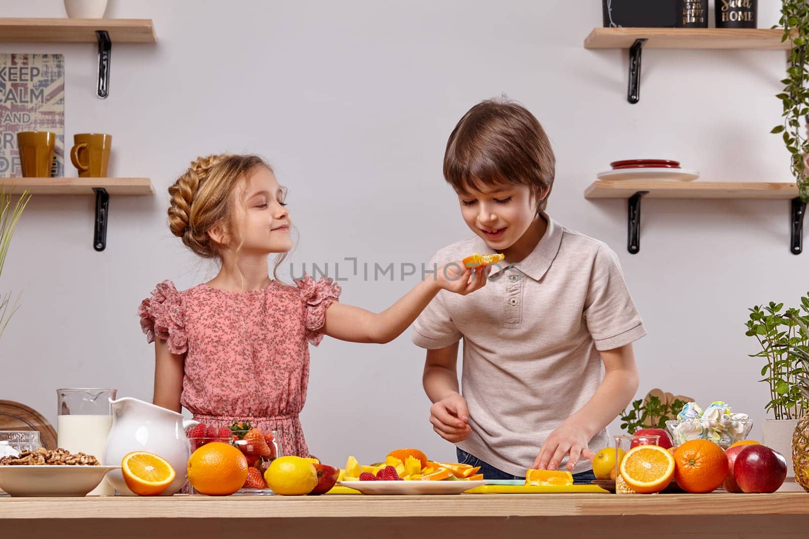 Cute cook couple. Nice boy with brown hair dressed in a light t-shirt and jeans with a cheerful little girl dressed in a pink dress with a braid in her hairstyle are at a kitchen against a white wall with shelves on it. Girl is treating a boy with some orange.