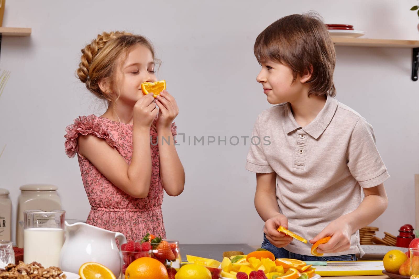 Cute cook couple. Handsome boy with brown hair dressed in a light t-shirt and jeans with a pretty little girl dressed in a pink dress with a braid in her hairstyle are at a kitchen against a white wall with shelves on it. Girl is smelling an orange and boy is looking at her.