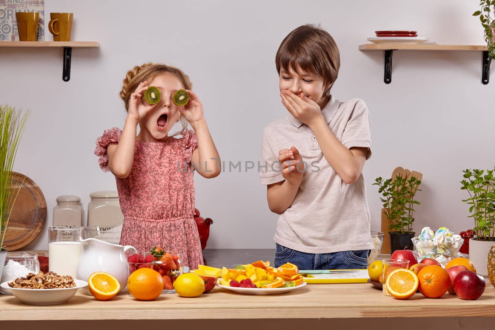 Cute cook couple. Boy with brown hair dressed in a light t-shirt and jeans with a little girl dressed in a pink dress with a braid in her hairstyle are at a kitchen against a white wall with shelves on it. Girl is holding a kiwi as if it were her eyes and opened her mouth widely and boy is laughing at her.
