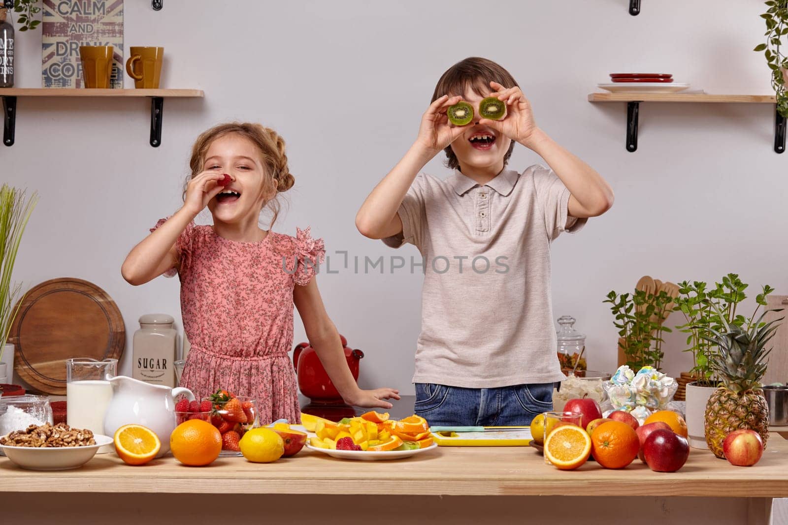 Cute cook couple. Handsome brunette boy dressed in a light t-shirt and jeans with a little blond girl dressed in a pink dress with a braid in her hairstyle are smiling at a kitchen against a white wall with shelves on it. Boy is holding a kiwi as if it were his eyes and girl is holding a strawberry as if it were her nose.