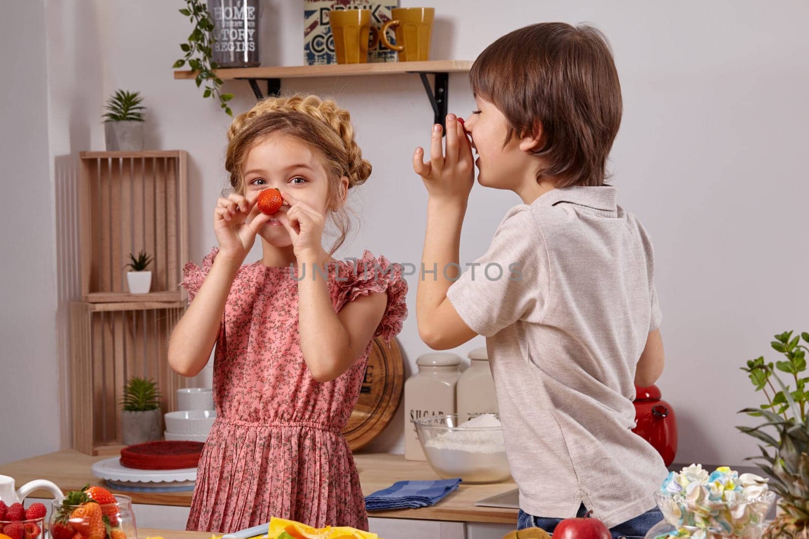 Cute cook couple. Brunette boy dressed in a light t-shirt and jeans with a little blond girl dressed in a pink dress with a braid in her hairstyle are smiling at a kitchen against a white wall with shelves on it. They are holding a strawberry as if it were their noses and laughing.