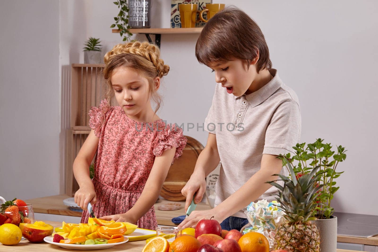 Cute cook couple. Little boy with brown hair dressed in a light t-shirt and jeans and a beautiful little girl with a braid in her hairstyle, dressed in a pink dress are cutting fruits together at a kitchen against a white wall with shelves on it.