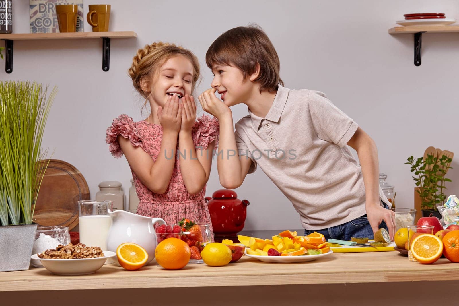 Cute cook couple. Pretty boy with brown hair dressed in a light t-shirt and jeans with a good-looking little girl dressed in a pink dress with a braid in her hairstyle are at a kitchen against a white wall with shelves on it. Boy is whispering something in a girl ear and she is laughing.