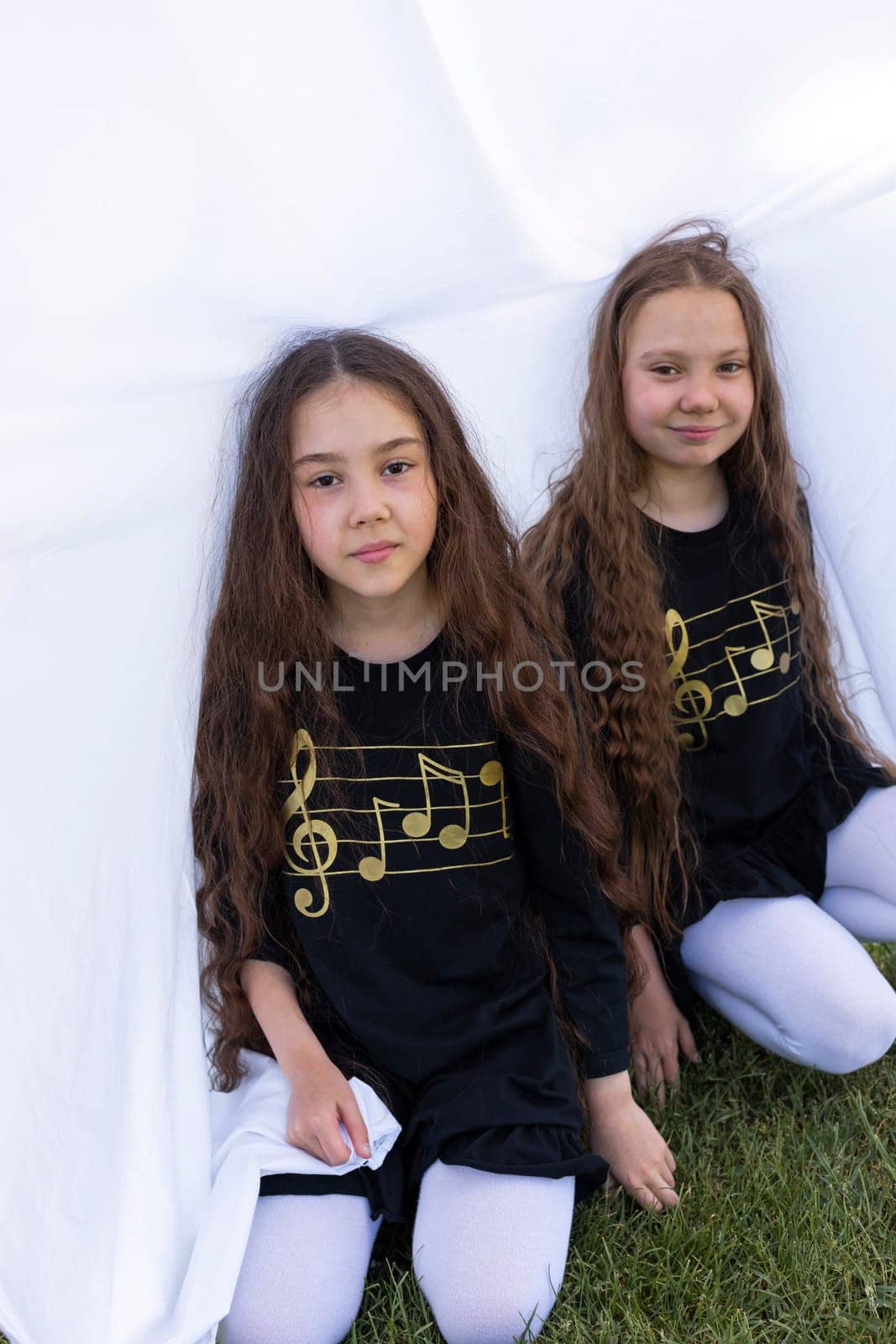 Two Little Girls, Sisters With Long Hair Having Fun Under Sheet On Grass in Park, Making Tent. Caucasian Asian Siblings Enjoy Time Together. Family Love And Care, True Friendship. Vertical Plane