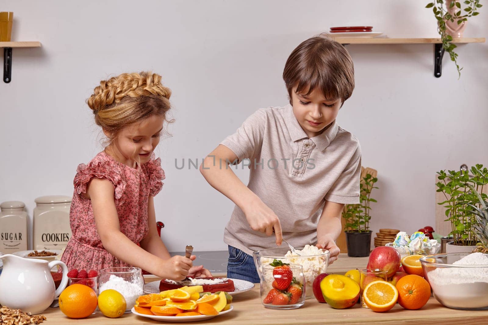 Pretty brunette boy dressed in a light t-shirt and jeans and a charming girl with a braid in her hair, wearing in a pink dress are making a cake at a kitchen, against a white wall with shelves on it. Girl is spreading some cottage cheese on a cake layer and boy is taking more cheese from a bowl.