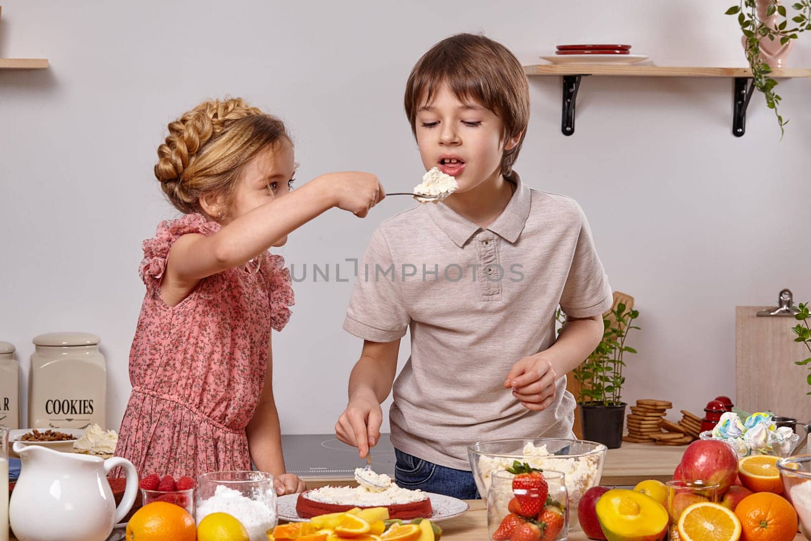 Brunette boy dressed in a light t-shirt and jeans and a girl with a braid in her hair, wearing in a pink dress are making a cake at a kitchen, against a white wall with shelves on it. Girl is treating a boy with some cottage cheese.