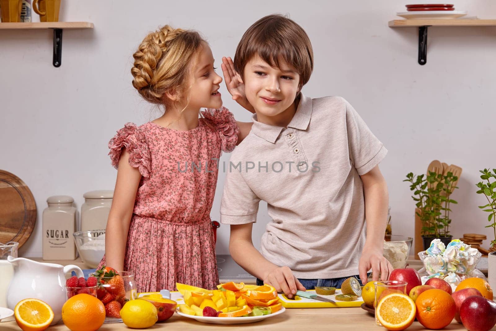 Cute kids are cooking together in a kitchen against a white wall with shelves on it. by nazarovsergey