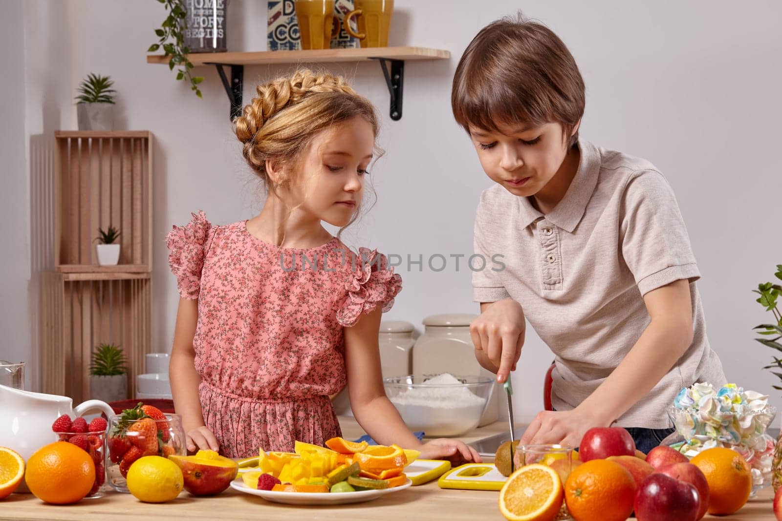 Cute cook couple. Boy with brown hair dressed in a light t-shirt and jeans with a little girl dressed in a pink dress with a braid in her hairstyle are at a kitchen against a white wall with shelves on it. Boy is cutting a kiwi and girl is looking at it.