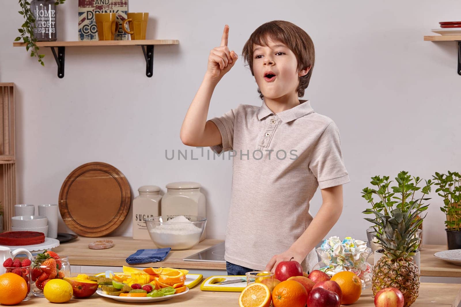 Cute little boy with brown hair is cooking at a kitchen against a white wall with shelves on it. by nazarovsergey