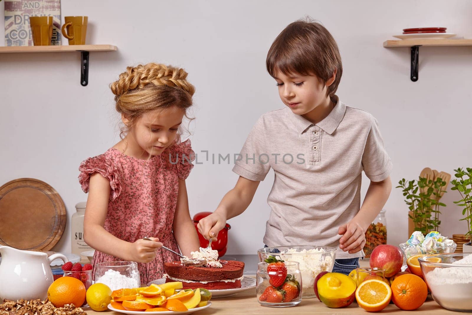 Brunette boy dressed in a light t-shirt and jeans and a cute girl with a braid in her hair, wearing in a pink dress are making a cake at a kitchen, against a white wall with shelves on it. They are spreading some cottage cheese on a cake layer.