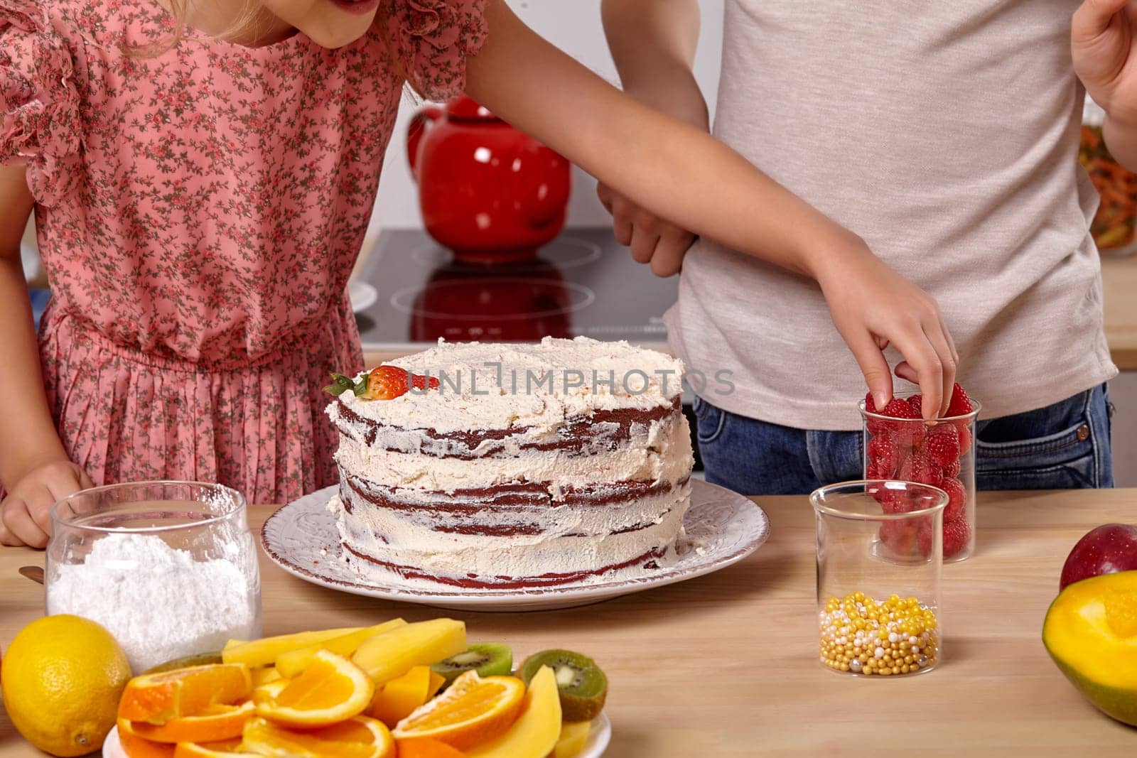 Close-up shot of a boy dressed in a light t-shirt and jeans and a girl wearing in a pink dress are making a cake at a kitchen. Girl is taking some raspberry from a glass.