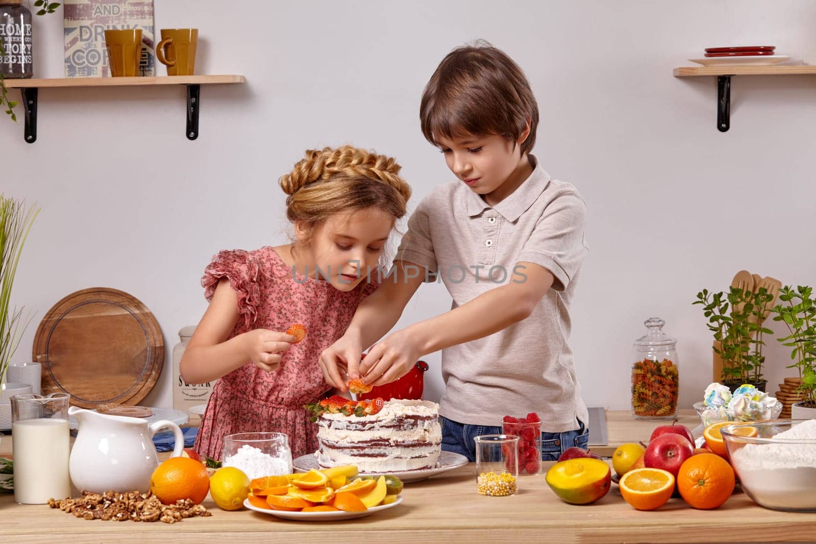 Brunette kid dressed in a light t-shirt and jeans and a beautiful girl with a braid in her hair, wearing in a pink dress are making a cake at a kitchen, against a white wall with shelves on it. They are decorating it with some strawberries.