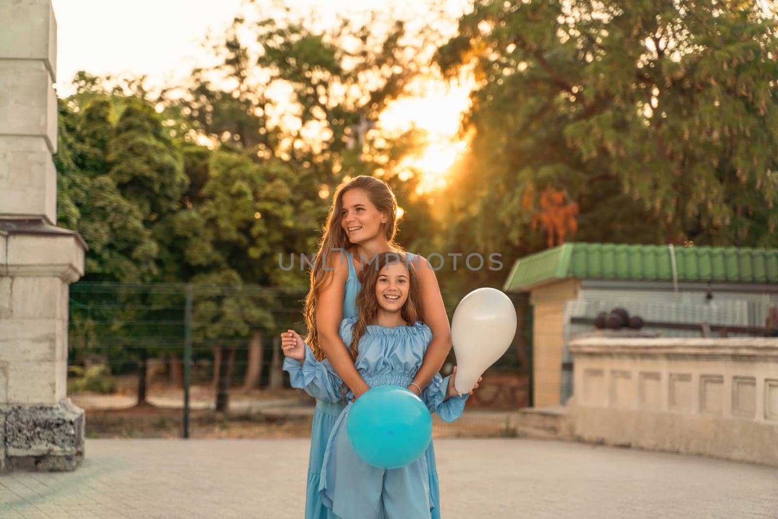 Mother daughter family sunset. Portrait of mother and daughter in blue dresses with flowing long hair against the backdrop of sunset. A woman hugs and presses the girl to her, holding balloons in her hands by Matiunina