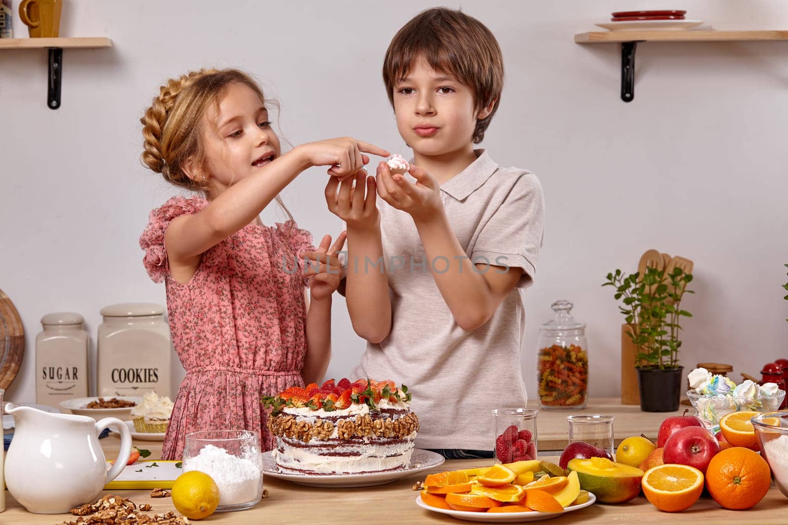 Little boy dressed in a light t-shirt and jeans and a wonderful girl with a braid in her hair, wearing in a pink dress are making a cake at a kitchen, against a white wall with shelves on it. Boy is holding a marshmallow in his hand and girl is looking at it.