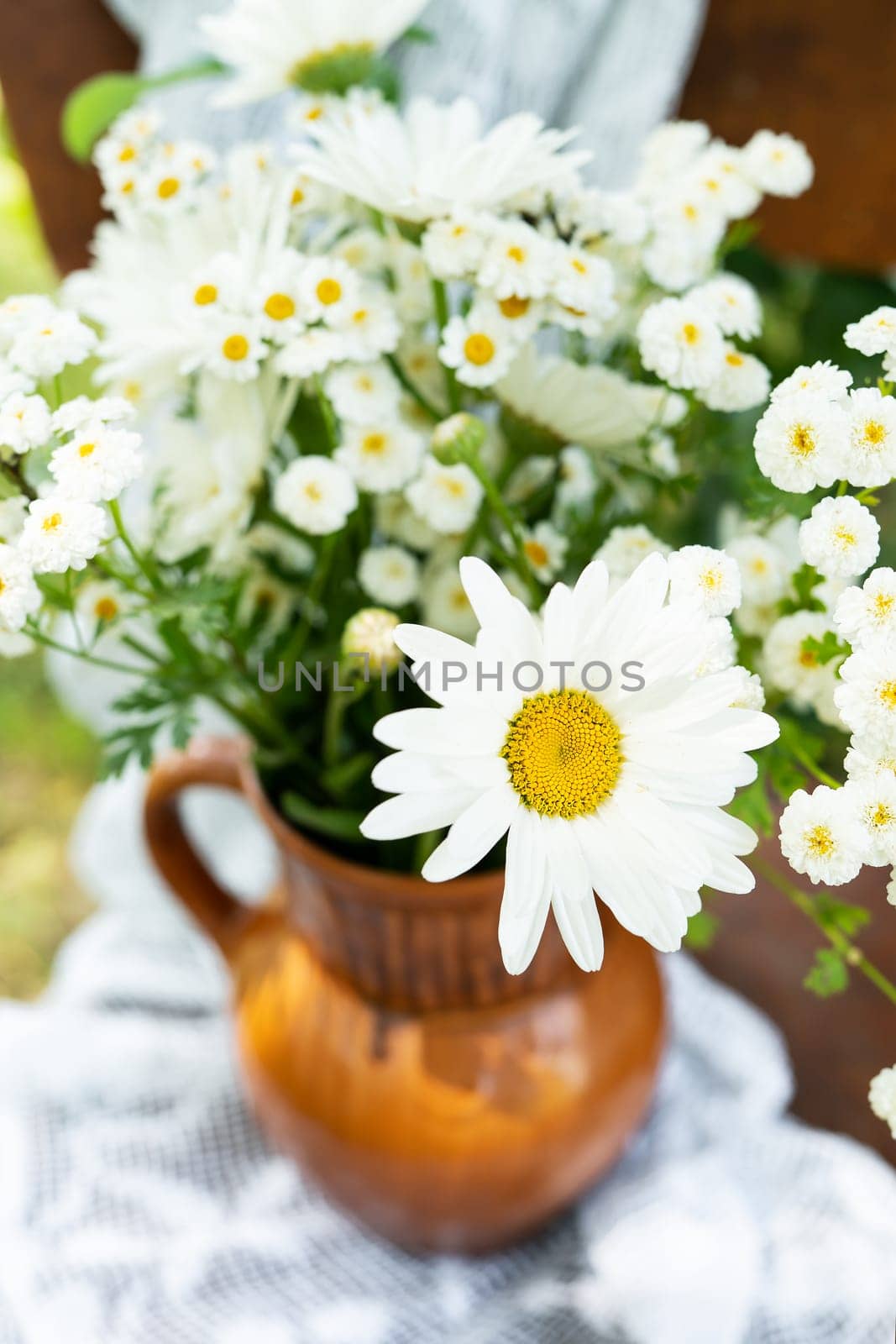 Beautiful chamomile flowers on a wooden old chair along with a lace doily on a green garden background. Summer atmosphere, simple home decor in the countryside. Vertical photo