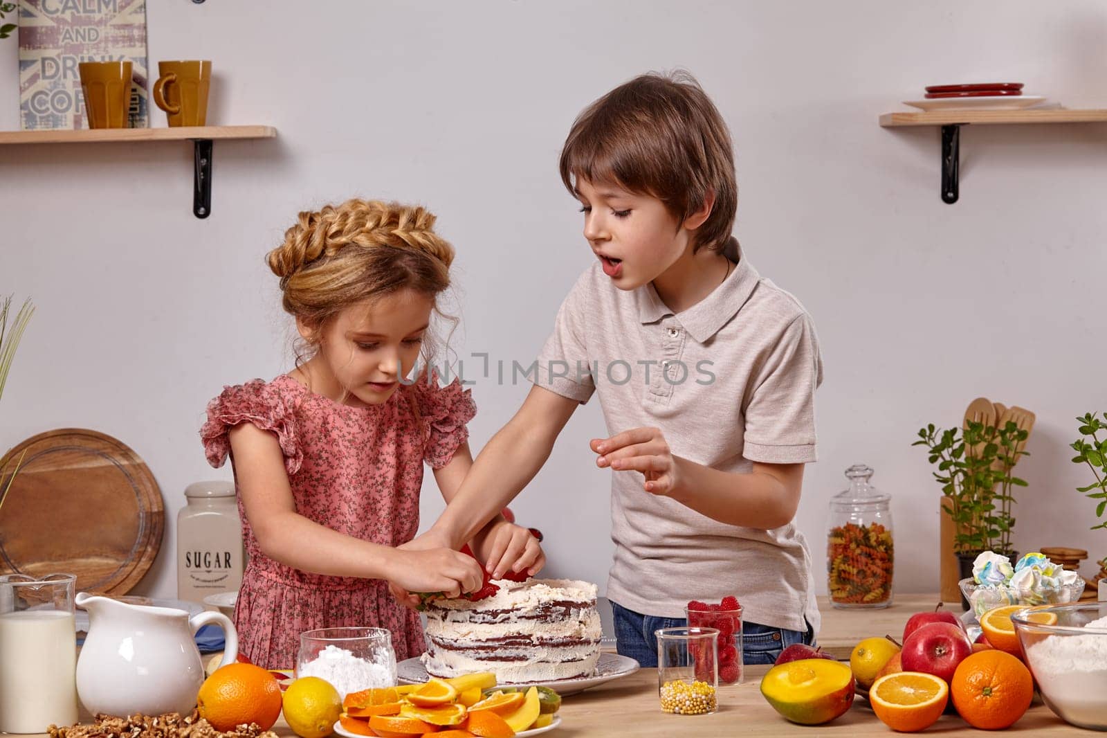 Little brunette kid dressed in a light t-shirt and jeans and a beautiful girl with a braid in her hair, wearing in a pink dress are making a cake at a kitchen, against a white wall with shelves on it. They are decorating a cake with some strawberries.