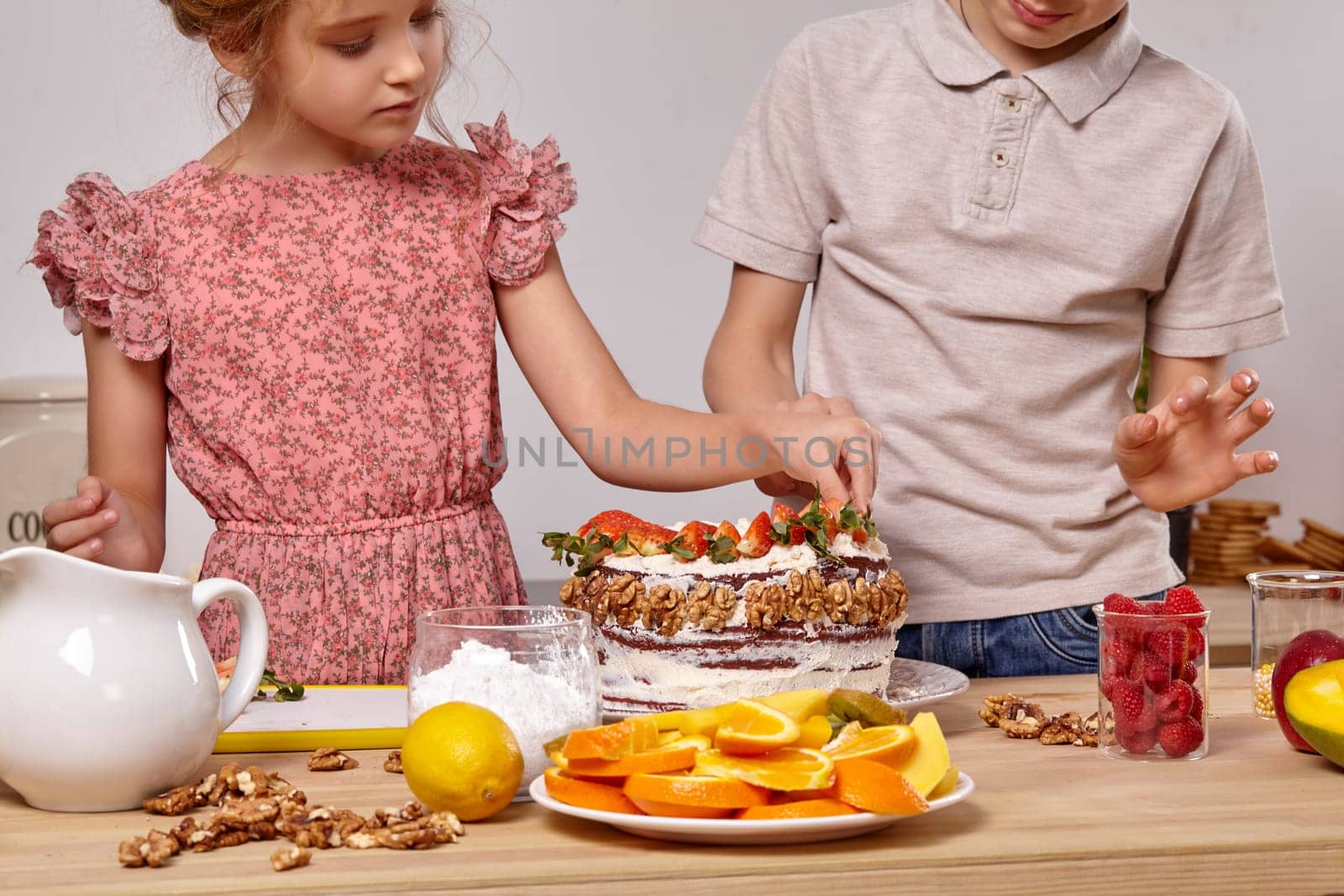 Pretty child dressed in a light t-shirt and jeans and a charming girl with a braid in her hair, wearing in a pink dress are making a cake at a kitchen, against a white wall. They are decorating it with a strawberry.