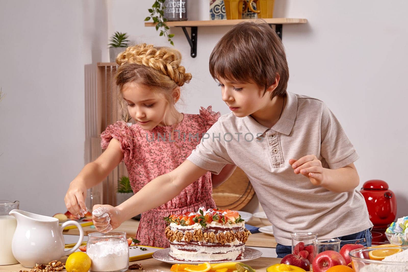 Little brunette boy dressed in a light t-shirt and jeans and a beautiful girl with a braid in her hair, wearing in a pink dress are making a cake at a kitchen, against a white wall with shelves on it. Children taking some powdered sugar in order to sprinkle a cake.