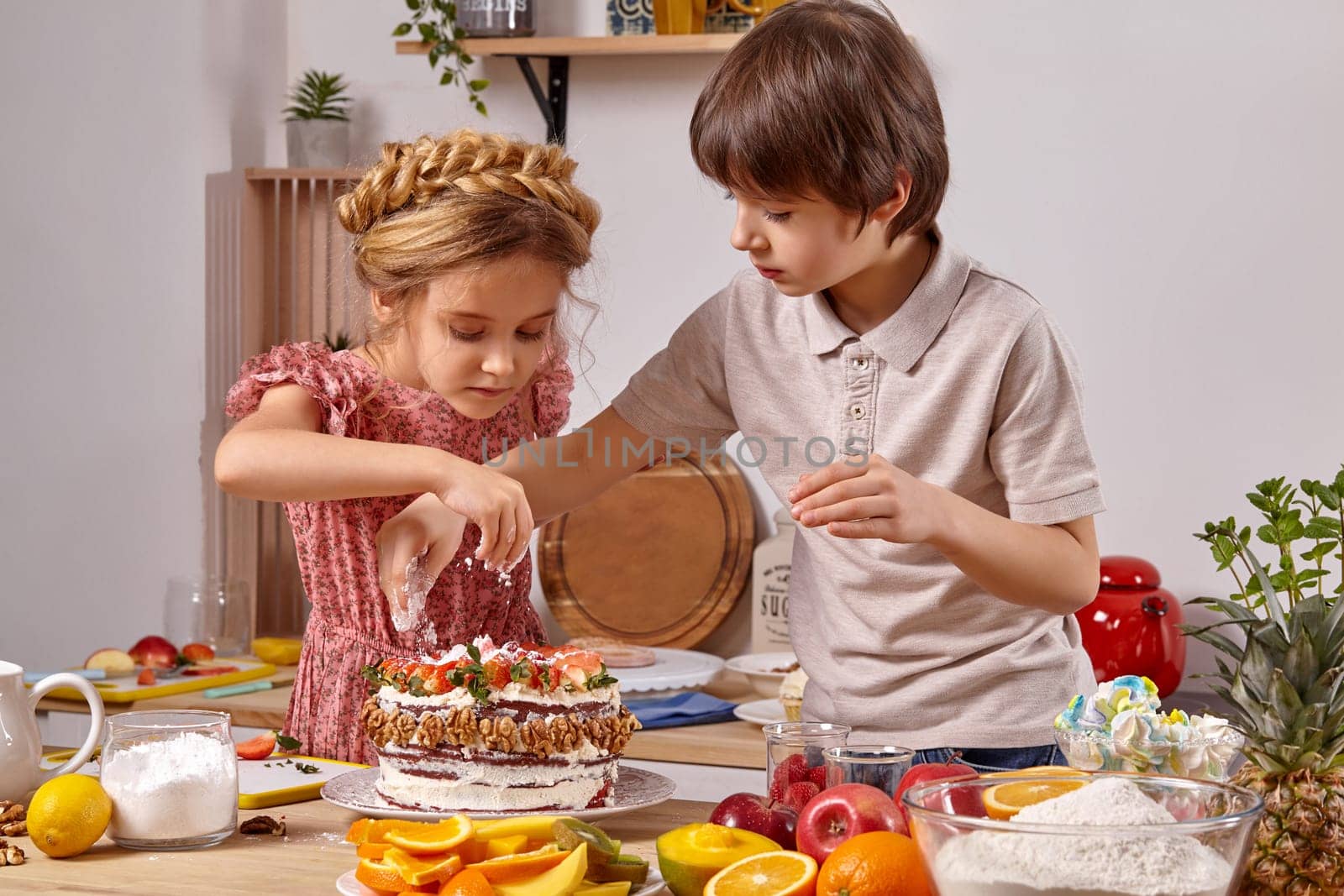 Kid dressed in a light t-shirt and jeans and a girl with a braid in her hair, wearing in a pink dress are making a cake at a kitchen, against a white wall with shelves on it. Children carefully sprinkle it with some powdered sugar.