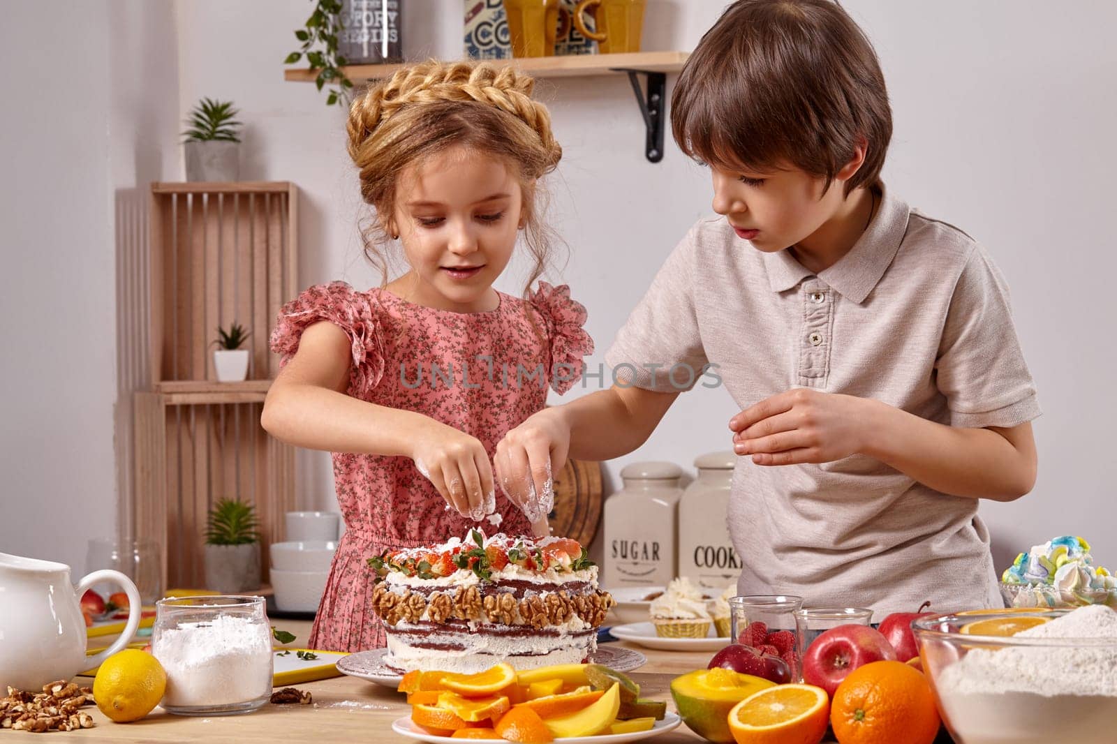 Brunette kid dressed in a light t-shirt and jeans and a blond girl with a braid in her hair, wearing in a pink dress are making a cake at a kitchen, against a white wall with shelves on it. Children carefully sprinkle it with some powdered sugar.