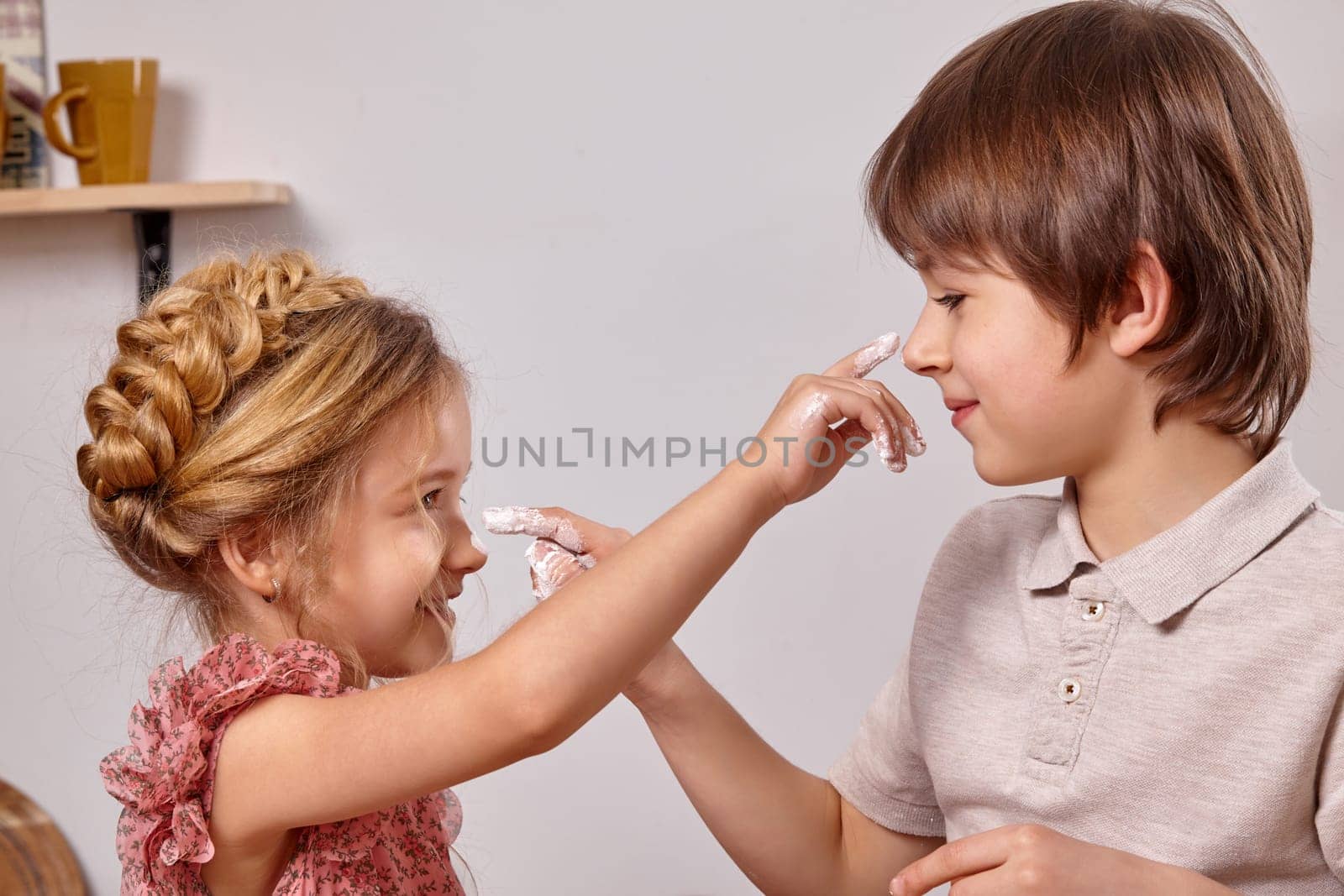 Funny boy dressed in a light t-shirt and jeans and a pretty girl with a braid in her hair, wearing in a pink dress are making a cake at a kitchen, against a white wall with shelves on it. They are smearing noses to each other with an icing sugar.