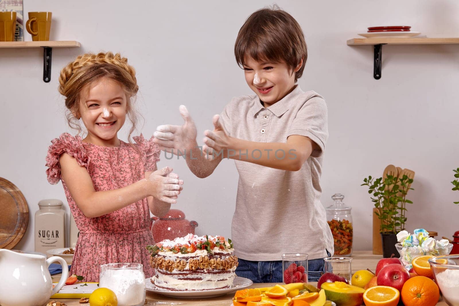 Brunette boy dressed in a light t-shirt and jeans and a little girl with a braid in her hair, wearing in a pink dress are making a cake at a kitchen, against a white wall with shelves on it. Children smeared their hands with powdered sugar and clapping it.