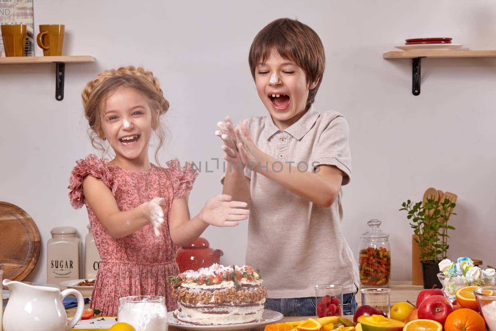 Little friends are making a cake together at a kitchen against a white wall with shelves on it. by nazarovsergey