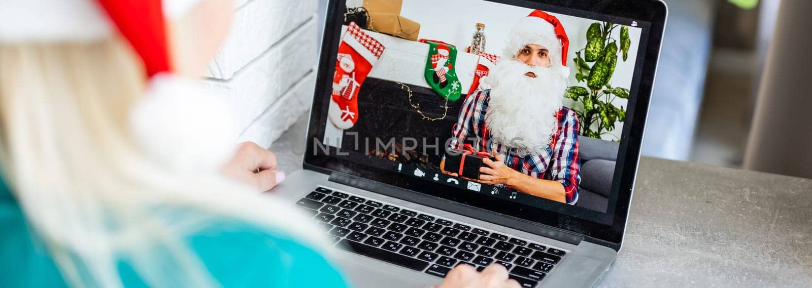 Smiling girl wearing Santa hat having video calling family by webcam. Woman with laptop sitting on kitchen at home using virtual meeting chat on holidays. Happy Christmas and New Year new normal. by Andelov13