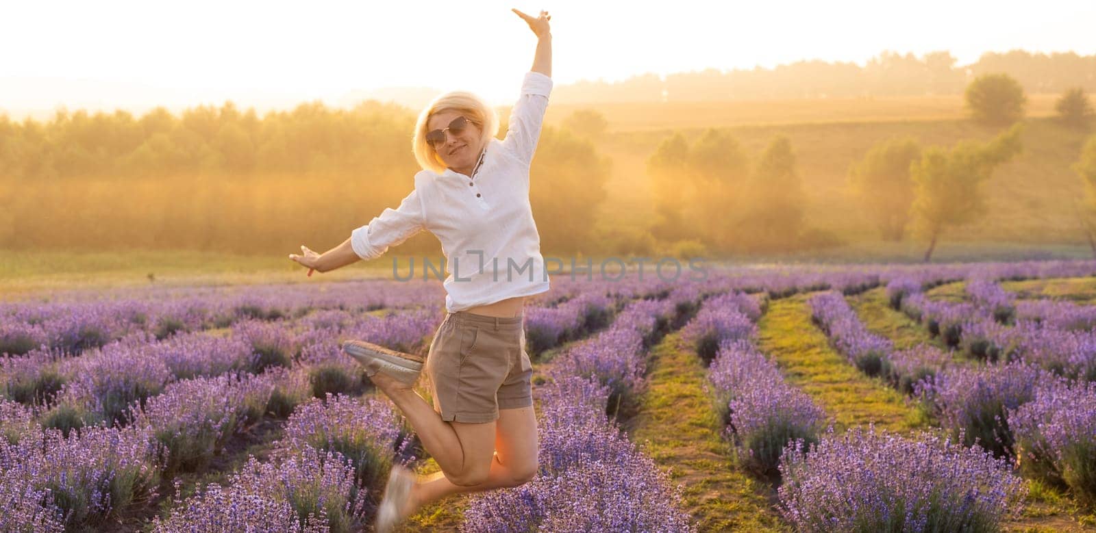 Beautiful young healthy woman with a white dress running joyfully through a lavender field holding a straw hat under the rays of the setting sun.