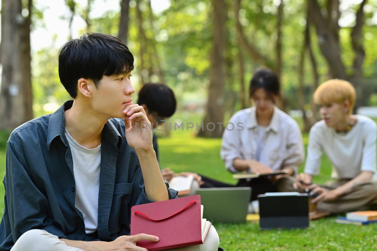 Thoughtful asian man student sitting on green grass in front of university building. Education, technology and lifestyle concept.