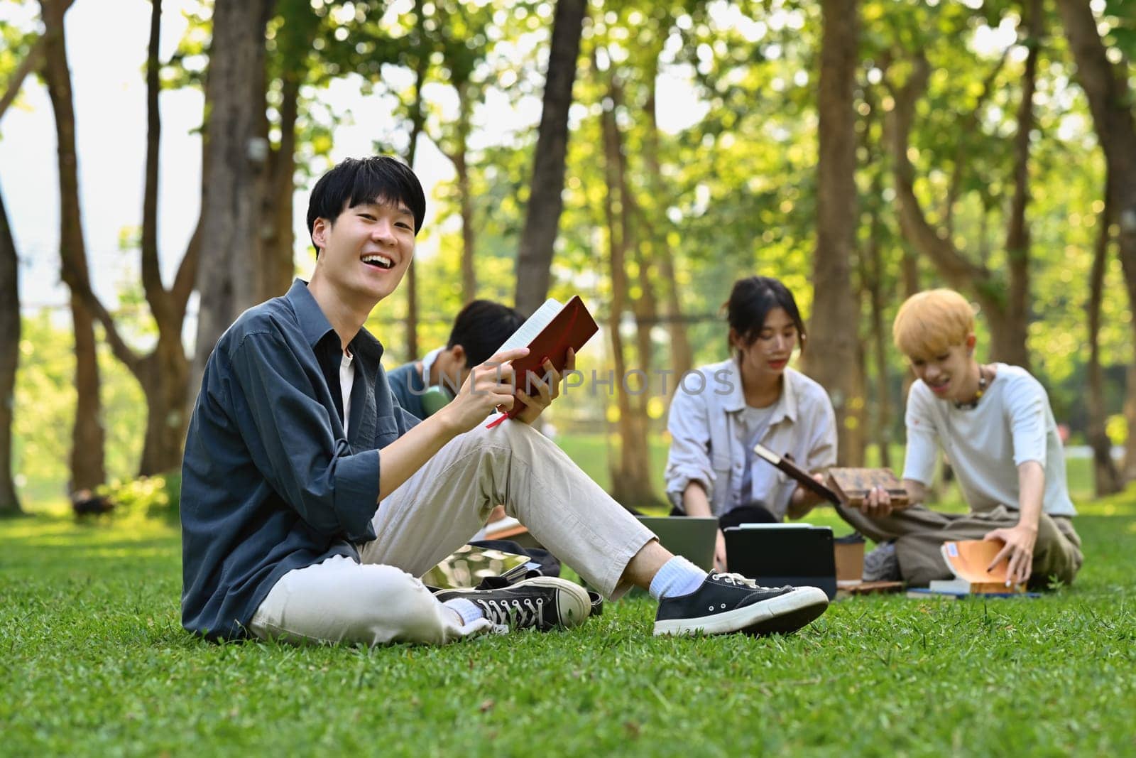 Smiling asian student man reading book on green grass in campus with friends sitting on background. Education and lifestyle concept.